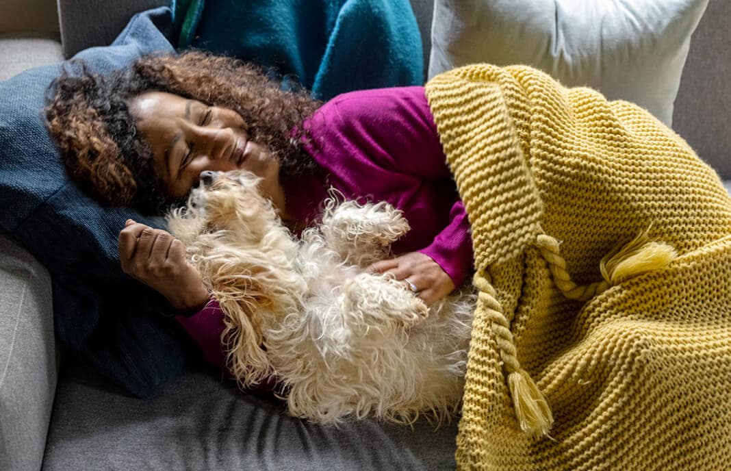 Woman in yellow blanket cuddling small dog on couch